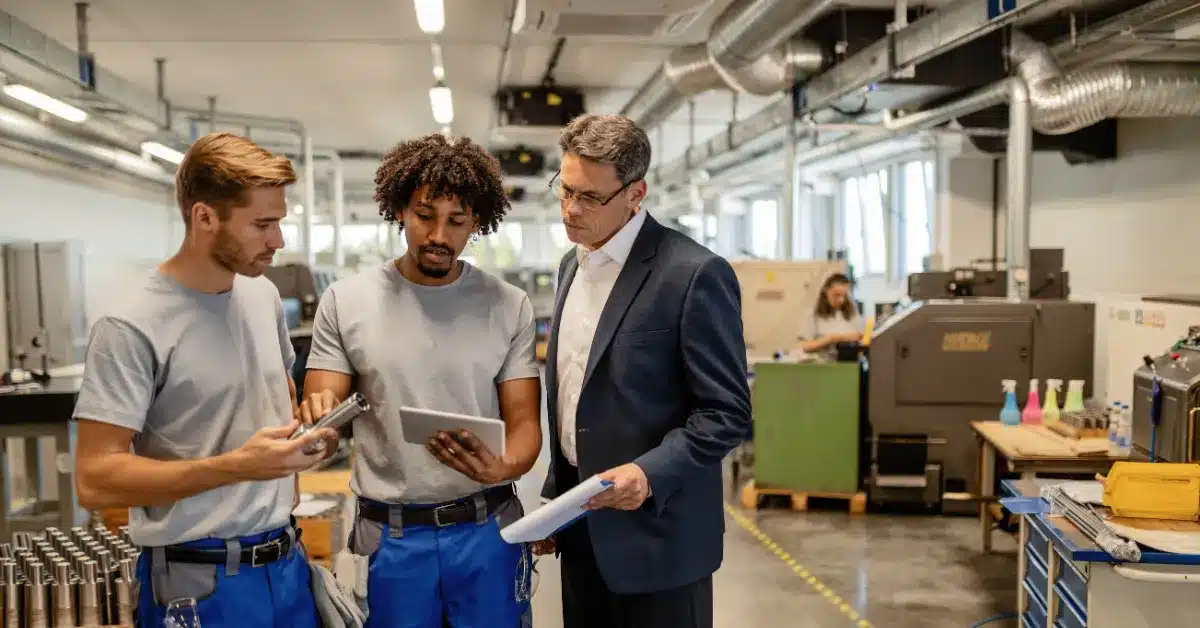 Two factory workers review a part with a supervisor in a suit inside a modern manufacturing facility, using a tablet and documents.