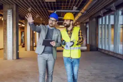 Project manager in a suit and blue hard hat discussing a build with a worker in a yellow hard hat and safety vest, walking through an unfinished commercial space.