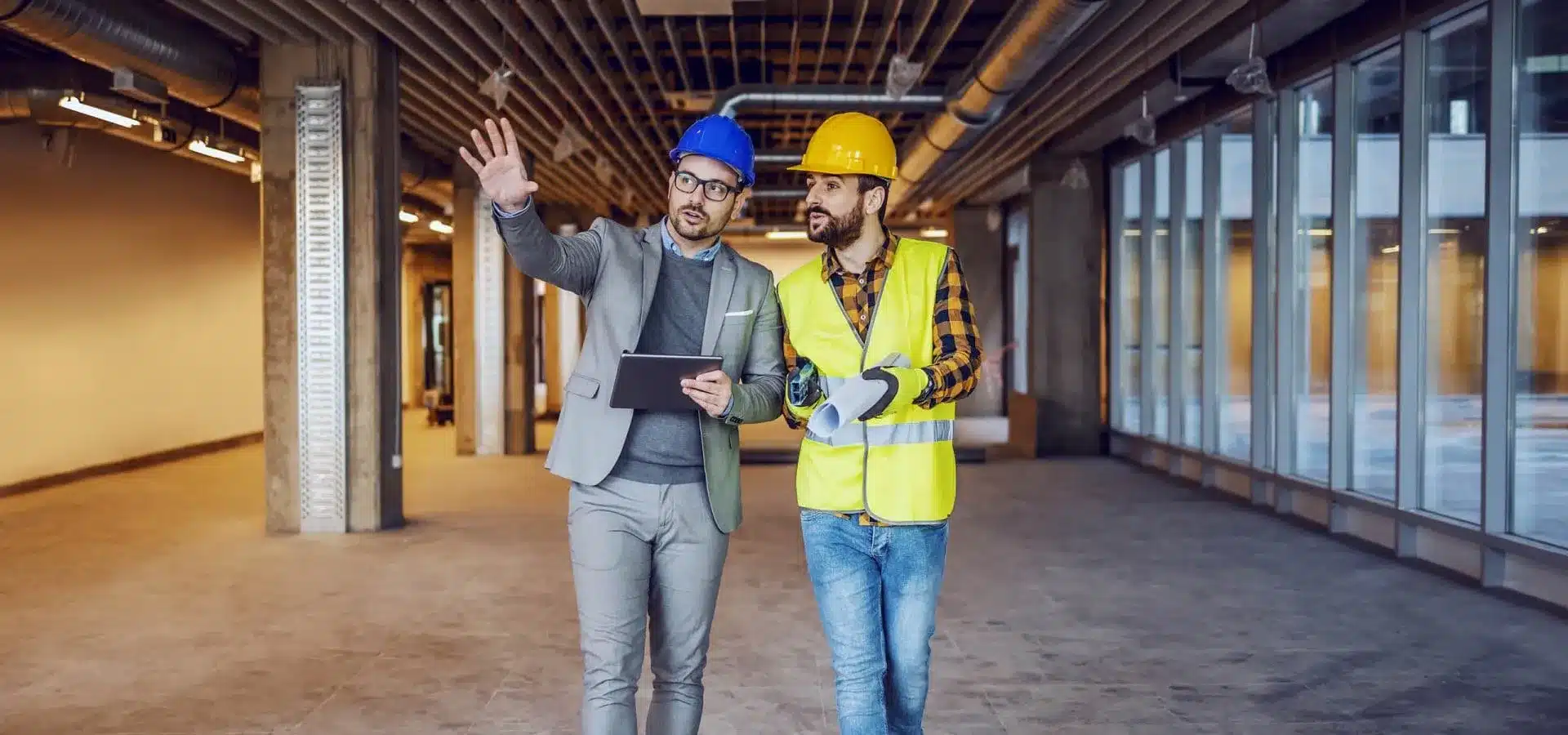 Project manager in a suit and blue hard hat discussing a build with a worker in a yellow hard hat and safety vest, walking through an unfinished commercial space.