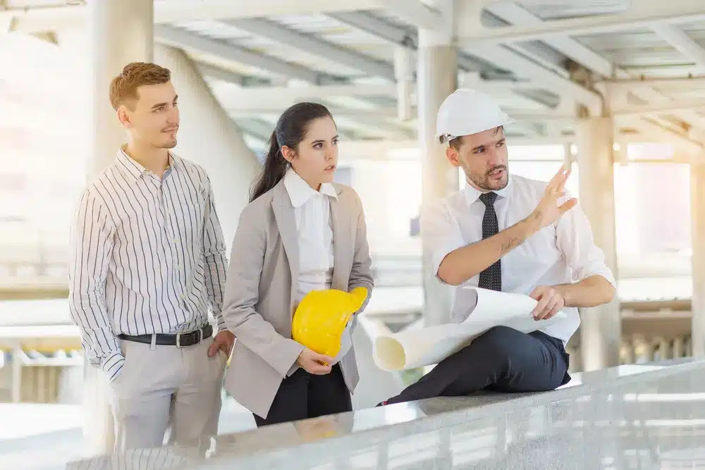 Construction team meeting: male supervisor in a hard hat gestures while holding blueprints, speaking to two colleagues; woman holds a yellow safety helmet.