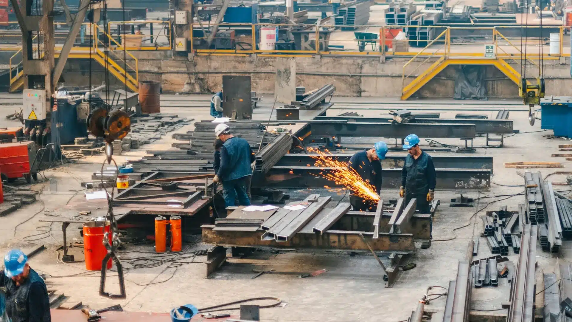Manufacturing workers in hard hats cutting and welding large steel beams inside an industrial workshop with sparks flying.