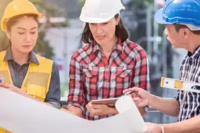 Three construction workers wearing safety helmets review blueprints and plans together at an outdoor job site.