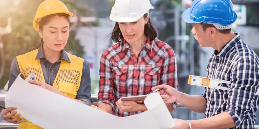 Three construction workers wearing safety helmets review blueprints and plans together at an outdoor job site.