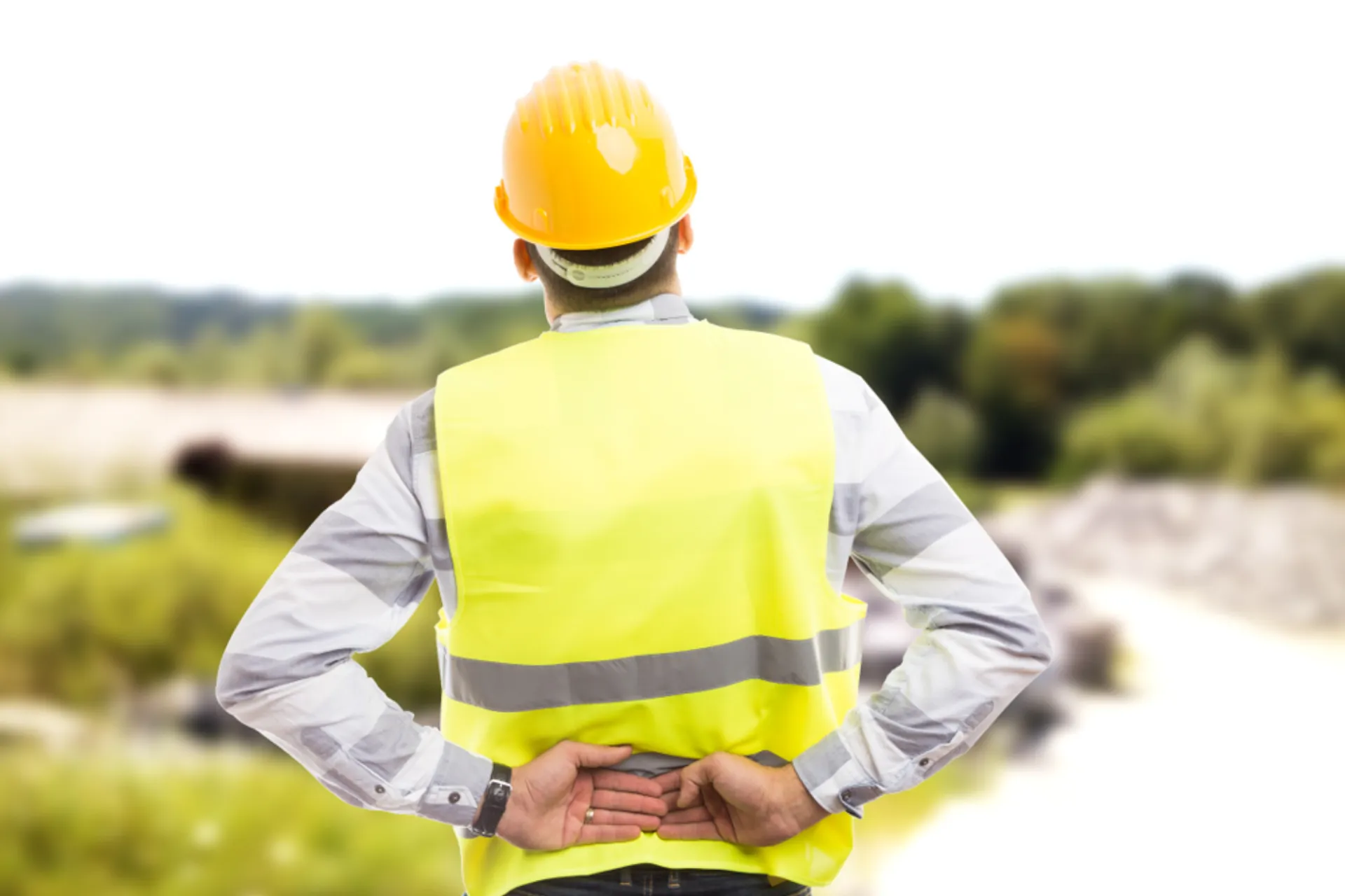 Construction supervisor in a hard hat and high-visibility vest stands with hands behind back, surveying a jobsite and planning next steps.