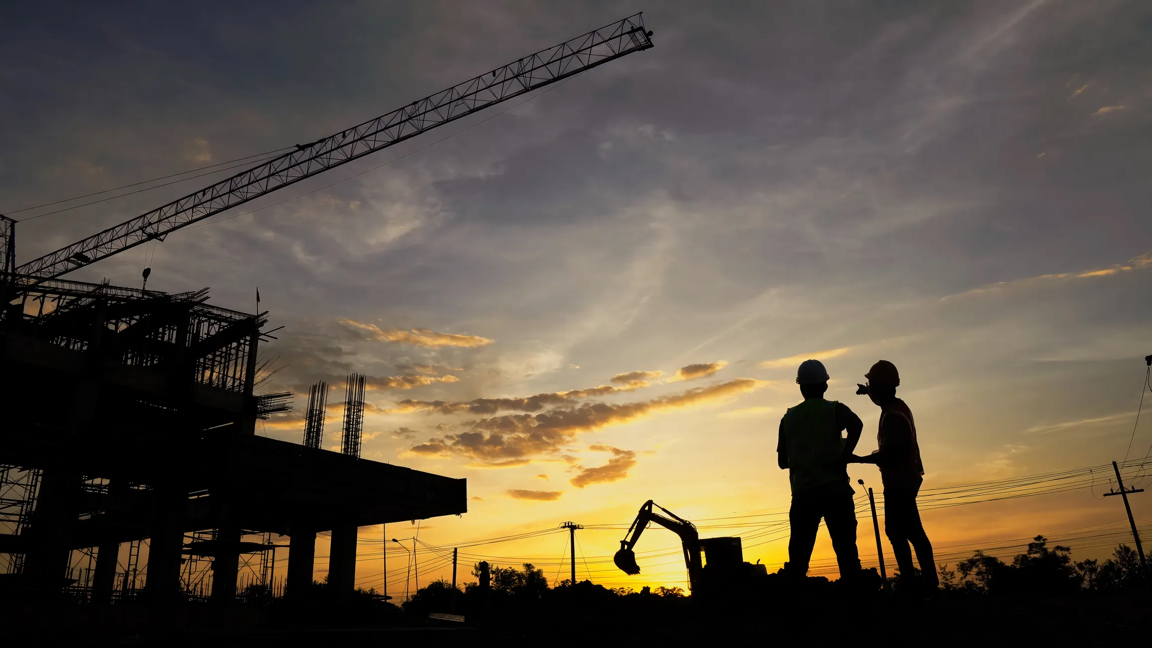 Two construction workers shown as black silhouettes standing on a construction site at sunset, with dramatic clouds filling the sky.