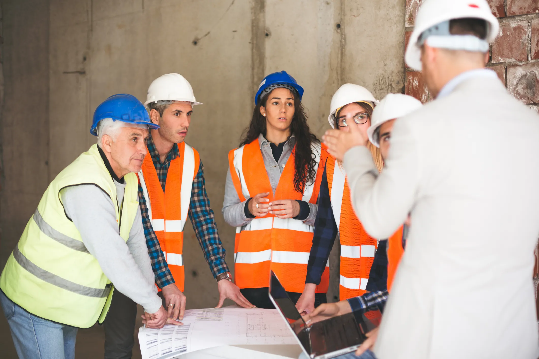 Group of construction workers, women and men wearing hard hats and safety vests, standing around a table attentively listening to a supervisor in a suit with his back to the camera, demonstrating effective team leadership on a construction site.